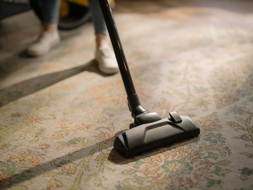 A person is kneeling on an ornate, patterned carpet in a domestic setting, using a yellow and black vacuum cleaner to perform surface cleaning. Their hands are placing a white cloth or cleaning access