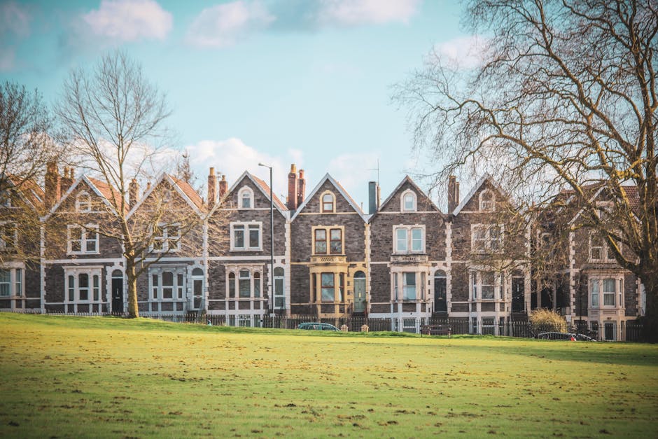 Image of a row of Victorian-style terraced houses made of grey stone with white window frames and gabled roofs, situated behind a well-maintained grassy park area with leafless trees. The houses feature multiple stories, large sash windows, and a central door with a small portico. The scene is brightly lit with natural daylight, and the surroundings appear clean and tidy, reflecting the property’s exterior maintenance. Cleaners Dulwich specializes in surface cleaning and sanitisation of residential exteriors, ensuring properties like these are kept in excellent condition and regularly deep cleaned to maintain hygiene and curb appeal.